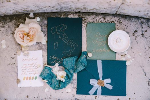 Blue mockup of invitations to wedding, blue envelope with seal print of sealing wax, a silk blue ribbon, a white sheet with the colors of dess code, pink flower against background of marble stone