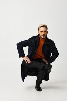 a blond man in glasses and in fashionable clothes sits on one leg and black boots. High quality photo