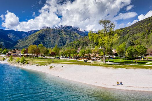 The Improbable aerial landscape of village Molveno, Italy, azure water of lake, The couple sits on a beach, snow covered mountains Dolomites on background, sunny weather, a piers