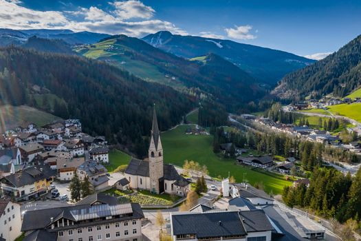 Aerial view of valley and small city with church, green slopes of the mountains of Italy, Trentino, San Martino in Badia, roofs of houses of settlements, green meadows, Dolomites on background, sunset