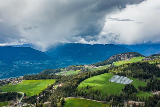 Aerial view of farm of solar batteries on green slopes of the mountains of Italy, Trentino, huge clouds over a valley, roofs of houses of settlements, green meadows, a clear energy, energy of the sun, drone view point