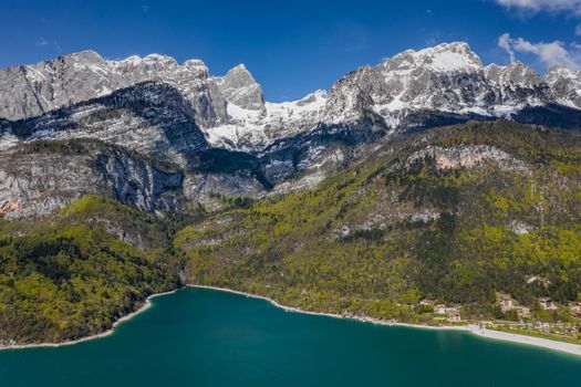 The Improbable aerial landscape of village Molveno, Italy, azure water of lake, empty beach, snow covered mountains Dolomites on background, roof top of chalet, sunny weather, a piers, coastline, slopes