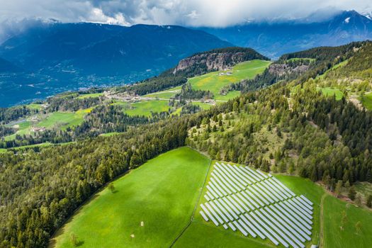Aerial view of farm of solar batteries on green slopes of the mountains of Italy, Trentino, huge clouds over a valley, roofs of houses of settlements, green meadows, a clear energy, energy of the sun, drone view point