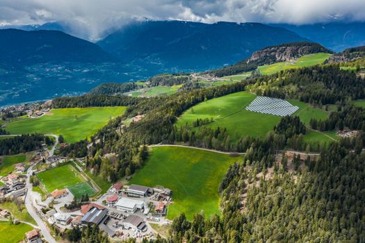 Aerial view of farm of solar batteries on green slopes of the mountains of Italy, Trentino, huge clouds over a valley, roofs of houses of settlements, green meadows, a clear energy, energy of the sun, drone view point