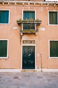 Two-storey peach-colored building with green wooden shutters for windows and door. A balcony above the front door with a forged metal grille and with flowers in pots on the railings.