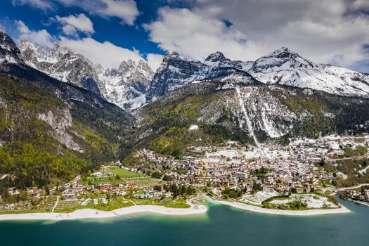 The Improbable aerial landscape of village Molveno, Italy, azure water of lake, empty beach, snow covered mountains Dolomites on background, roof top of chalet, sunny weather, a piers, coastline, slopes