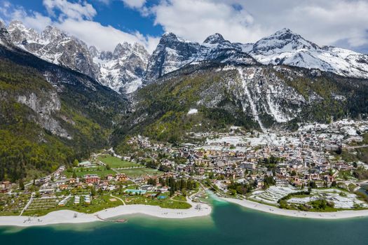 The Improbable aerial landscape of village Molveno, Italy, azure water of lake, empty beach, snow covered mountains Dolomites on background, roof top of chalet, sunny weather, a piers, coastline, slopes