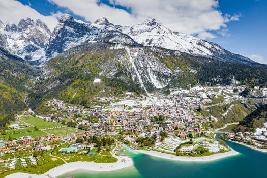 The Improbable aerial landscape of village Molveno, Italy, azure water of lake, empty beach, snow covered mountains Dolomites on background, roof top of chalet, sunny weather, a piers, coastline, slopes