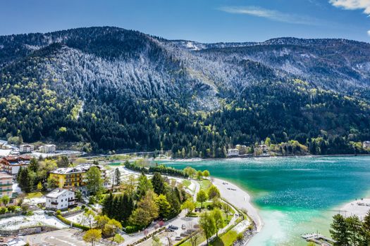 The Improbable aerial landscape of village Molveno, Italy, azure water of lake, empty beach, snow covered mountains Dolomites on background, roof top of chalet, sunny weather, a piers, coastline, slopes