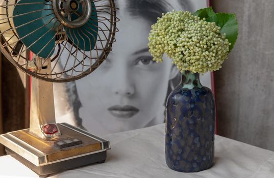 Bangkok, Thailand -Feb 06, 2021 : Bouquet of flowers in Blue ceramic vase and vintage fan on white textured table cloth in front of Classic chinese poster movie frame with old cement wall. Home decor, Selective focus.