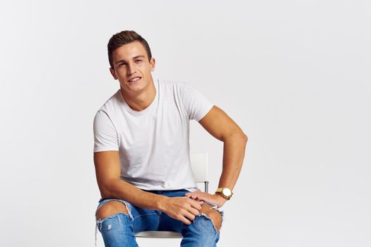 Man on a chair indoors torn jeans white t-shirt handsome face model light background. High quality photo