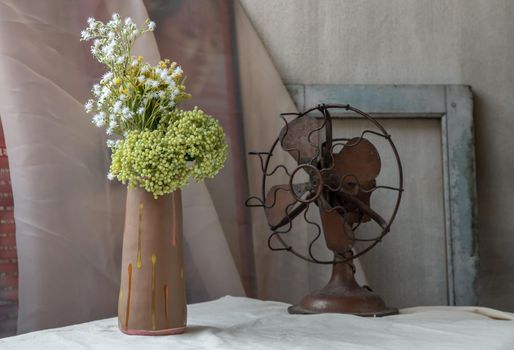 Bouquet of green and white flowers in Handmade ceramic vase and Vintage fan on white textured table cloth with old cement wall. Home decor, Selective focus.