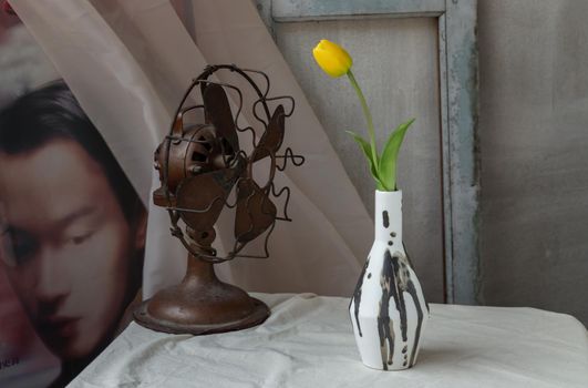 Bangkok, Thailand - Feb 06, 2021 : Yellow flower in Black and White Handmade ceramic vase and Vintage fan on white textured table cloth in front of Classic chinese poster movie frame with old cement wall. Home decor, Selective focus.