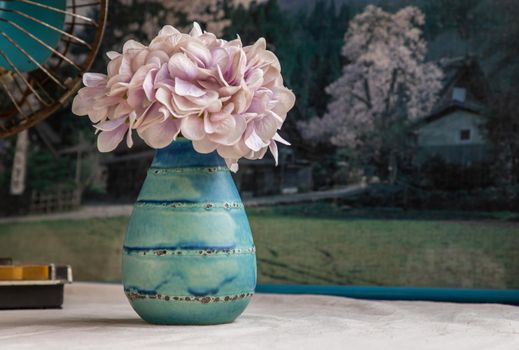 Bangkok, Thailand - Feb 06, 2021 : Bouquet of purple flowers in Blue handmade ceramic vase and old vintage fan on white textured table cloth in front of Beautiful scenery wallpaper. Home decor, Selective focus.