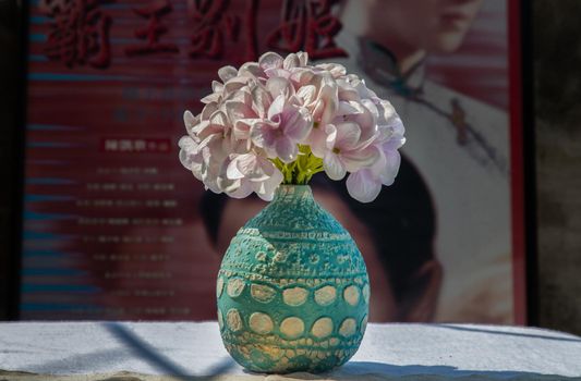 Bangkok, Thailand - Feb 06, 2021: Bouquet of purple flowers in Blue handmade ceramic vase on white textured table cloth in front of Classic chinese poster movie frame. Home decor, No focus, specifically.