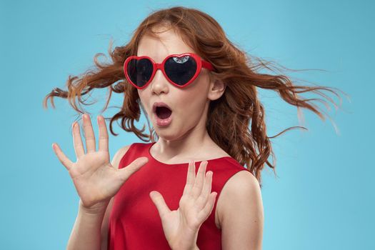 Little girl in sunglasses in the form of hearts red dress curly hair blue background. High quality photo