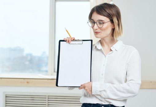 A woman is holding a folder with a white sheet of paper and a window in the background. High quality photo