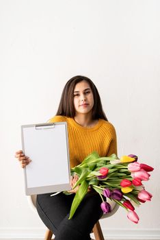 Beautiful young brunette woman in yellow sweater with bouquet of fresh pink tulips and blank clipboard for mock up