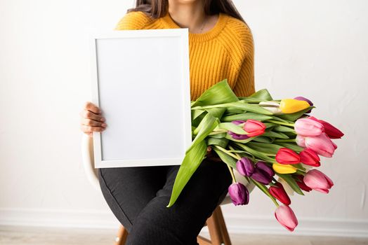 Beautiful young brunette woman in yellow sweater with bouquet of fresh pink tulips and blank frame for mock up