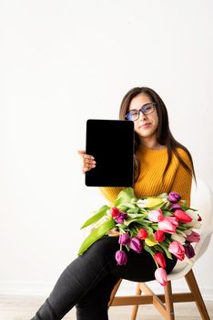 Beautiful young brunette woman in yellow sweater with bouquet of fresh pink tulips and blank digital tablet for mock up