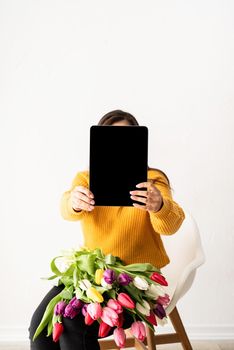 Beautiful young brunette woman in yellow sweater with bouquet of fresh pink tulips and blank digital tablet for mock up