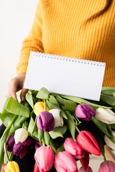 Beautiful young brunette woman in yellow sweater with bouquet of fresh pink tulips and blank calendar for mock up