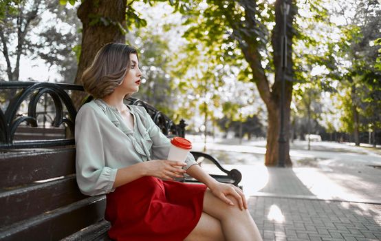 woman sits on a bench in the park in nature and hold a cup of coffee in her hand. High quality photo