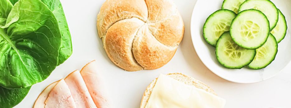 Food products and ingredients for making sandwich. Ham, cheese, burger bun, lettuce, cucumber and tomato as recipe flatlay on marble kitchen table background