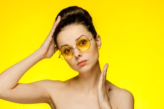 brunette with bare shoulders holds hands near face clear skin yellow glasses. High quality photo