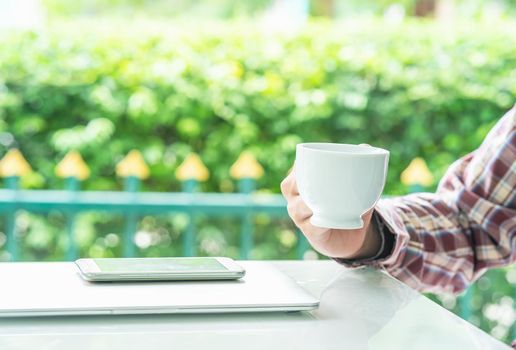 Male hands holding smart phone and cup of coffee on the table  outdoor with blurred green plant background