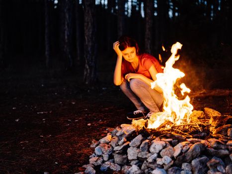 A woman sits near the fire and looks at the fire in a pine forest. High quality photo
