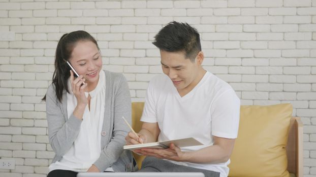 Happy Asian family couple husband and wife working confirming booking having talking call phone conversation and making writing notes of information together from home in living room. small business