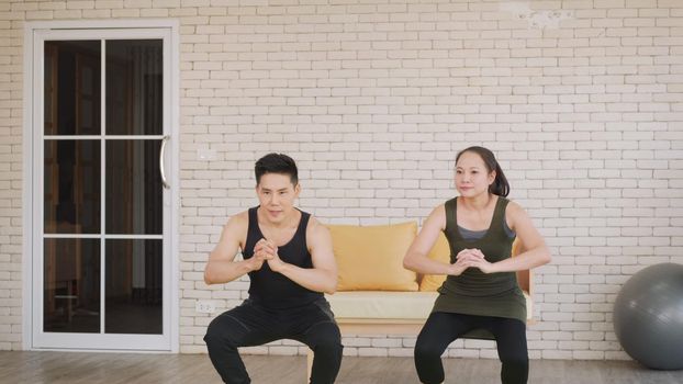 Happy Asian beautiful lifestyle family couple doing YOGA standing meditating on exercise sitting squat position workout at home together on mat. Two people sport healthy concept.