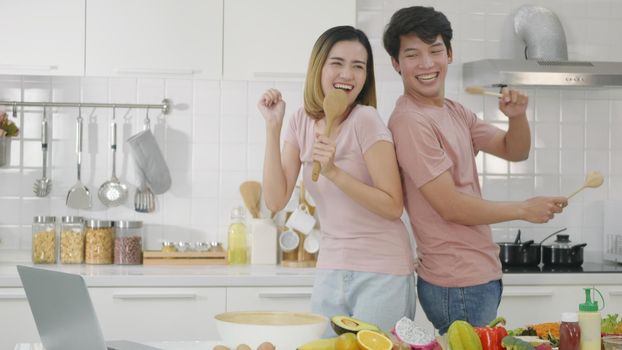 Happy Asian beautiful family couple husband and wife singing in kitchenware microphones in kitchen together having fun dance listen music at home. Two people dancing