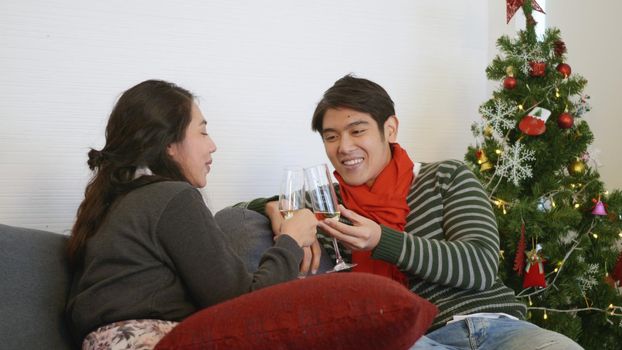 Happy Asian beautiful young family couple husband and wife on sofa in home living room, man and woman sit sofa in love clink glasses with champagne enjoy and drink winter holidays together