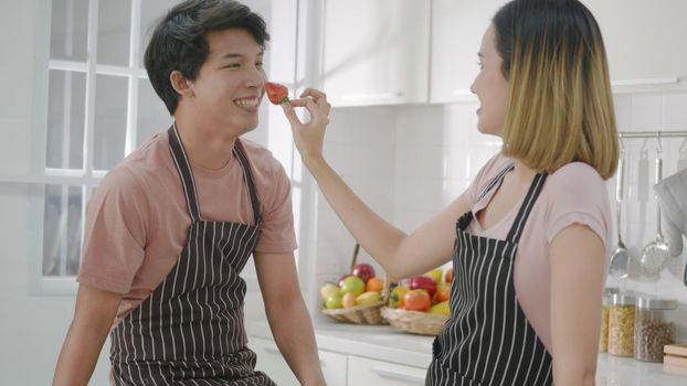 Happy Asian beautiful young family couple husband and wife enjoying smile and laugh, woman hold the strawberry to the man's nose spending time together in kitchen at home.