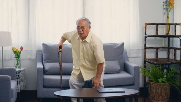 Old man grandfather with eyeglasses used a walking stick to lean the ground slowly stand up from the sofa carefully in living room at home. Asian senior old gray-haired suffering from knee pain ache