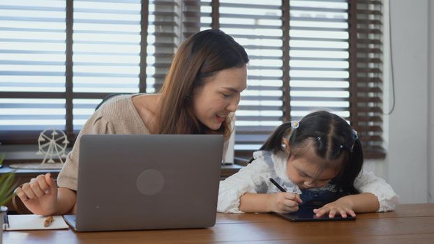 Asian young mother working with laptop computer at home and help daughter doing homework, Single mom freelancer work from home and helping his girl kid teaching homework on desk