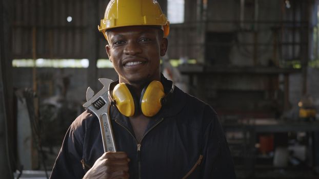 Portrait American industrial black young worker man smiling with helmet and ear protection in front machine, Engineer standing holding wrench on his shoulder at work in industry factory.