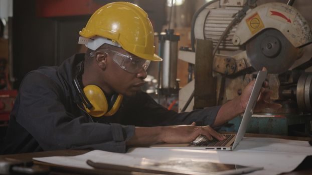 American industrial black young worker man with yellow helmet and ear protection typing keyboard of laptop computer in front machine, Confident engineer at work in the industry factory