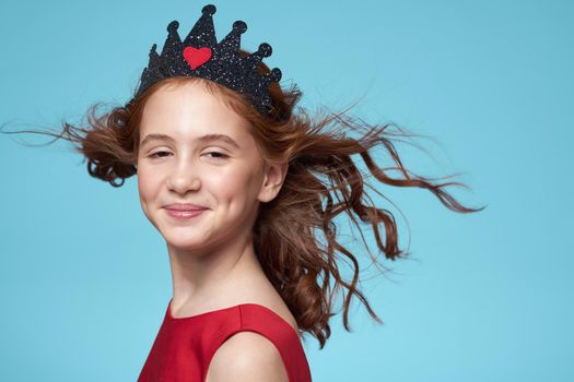 little girl with curly hair with a crown on her head red dress Smile blue background. High quality photo