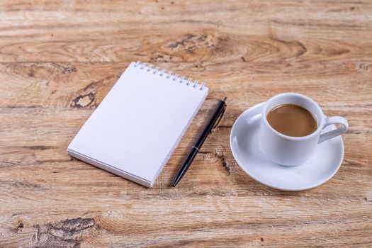 Small white coffee cup on a saucer stands on a wooden table next to a notebook and a pen, concept of coffee break at work, meeting, taking notes or planning