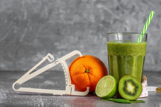 Fresh smoothie drink in a tall glass glass with centimeter wrapped around it and a caliper next to it, surrounded by fruits on a gray concrete background. The concept of proper nutrition, weight loss