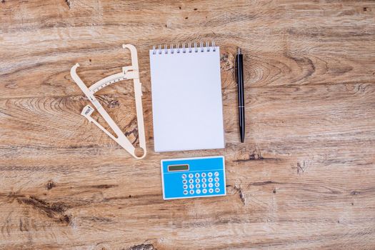 Blank sheet of notepad pen calculator and caliper on wooden table top view layout. The concept of proper nutrition, calorie counting, weight loss.