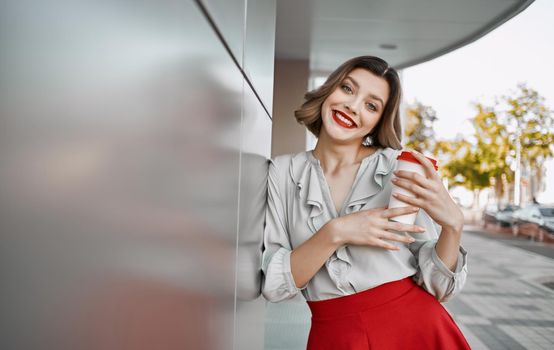 pretty blonde with a cup of coffee near the building on the street and red skirt white shirt. High quality photo