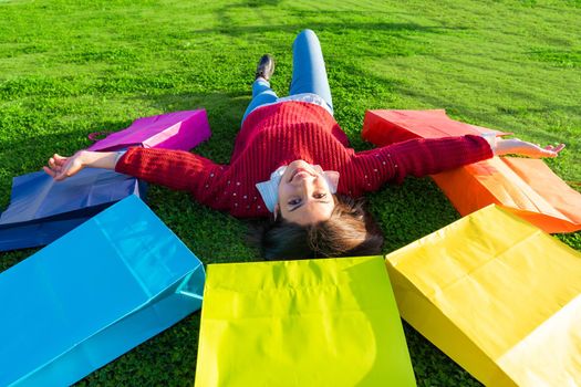 BEautiful happy girl lying on the grass of city park with open arms among seven shopping bags colored like iris wearing medical protection mask due to Coronavirus. New normal habits with Covid concept