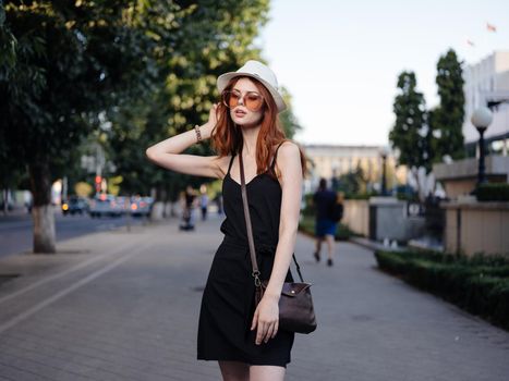 A woman in a black dress and in a white hat with a bag on her shoulder on the street. High quality photo