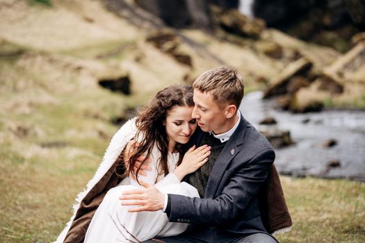 The wedding couple sits on the bank of a mountain river, at a table for a wedding dinner, made of wooden pallet, painted with moss and driftwood. The groom hugs the bride under a woolen plaid.