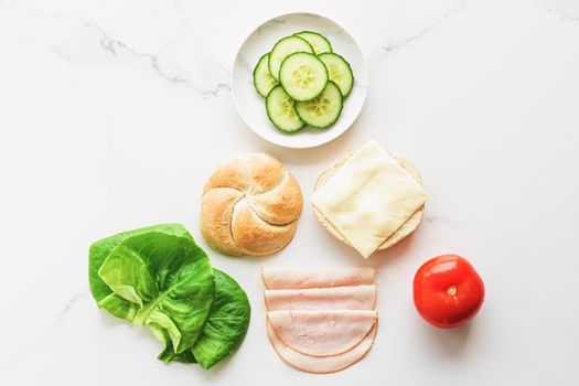 Food products and ingredients for making sandwich. Ham, cheese, burger bun, lettuce, cucumber and tomato as recipe flatlay on marble kitchen table background