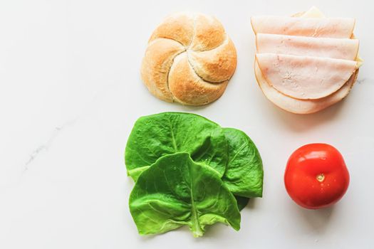 Food products and ingredients for making sandwich. Ham, cheese, burger bun, lettuce, cucumber and tomato as recipe flatlay on marble kitchen table background